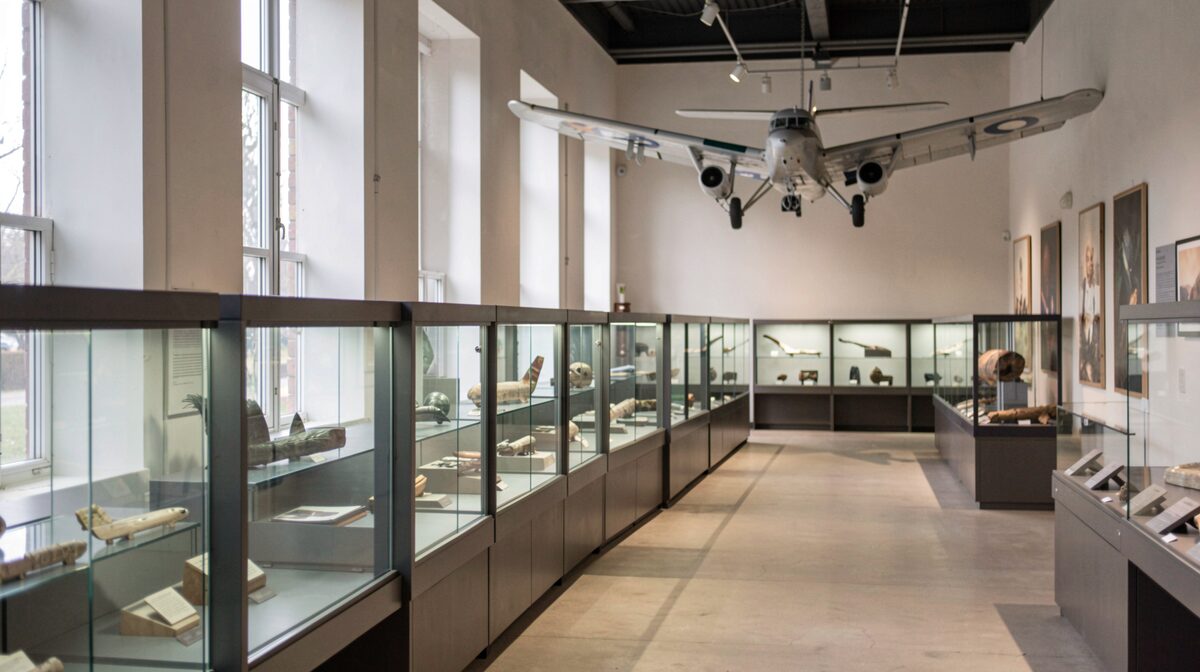 Vue d'une salle de musée local éclairée par une lumière naturelle latérale, vitrines vitrées alignées le long d'un mur, un avion léger suspendu au plafond en arrière-plan, cadrage large à hauteur d'œil.