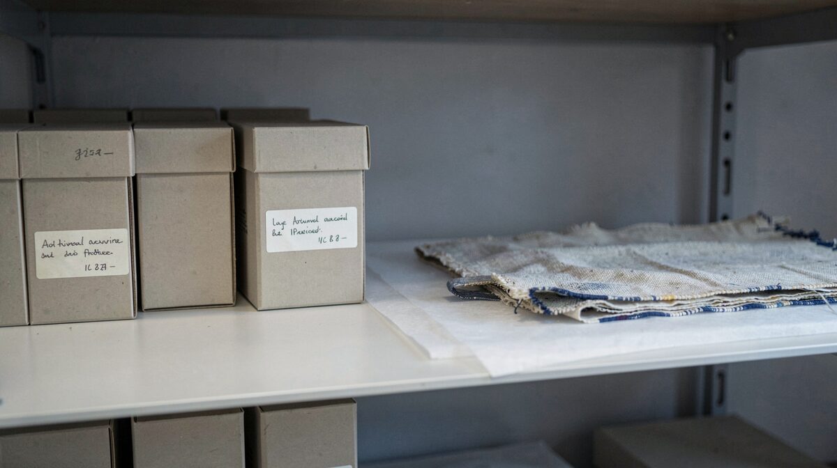 Une étagère de réserve de musée en lumière froide et diffuse, avec des boîtes d'archives neutres alignées, une étiquette manuscrite visible sur l'une d'elles et un objet textile posé à plat sur un support sans acide, cadrage frontal à hauteur de tablette.